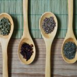 Overhead view of wooden spoons filled with spices and herbs on a bamboo mat, ideal for culinary themes.